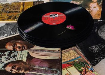 Artystyczna kompozycja ze stosem płyt winylowych i okładkami albumów z muzyką. Na wierzchniej płycie leży mały różowy kwiat