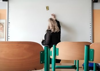 Nauczycielka w sali lekcyjnej przy białej tablicy, na pierwszym planie puste krzesła