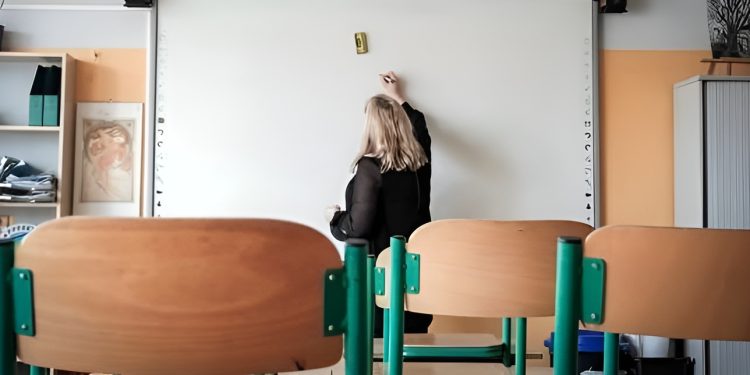 Nauczycielka w sali lekcyjnej przy białej tablicy, na pierwszym planie puste krzesła